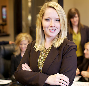 A woman smiles at the camera while standing with arms crossed in front of a group of coworkers
